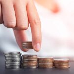 Hand placing a silver coin on a stack of coins, with several stacks of varying heights in the foreground.