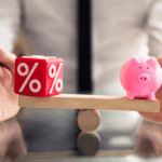 A wooden balance scale with a red percentage symbol block on one side and a pink piggy bank on the other, held by a person in a white shirt and black tie.