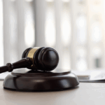 Brown wooden gavel resting on a circular base with a blurred background of light-colored vertical blinds.