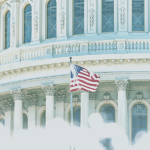 U.S. Capitol building with a prominently displayed American flag in front, surrounded by a soft, cloudy atmosphere.