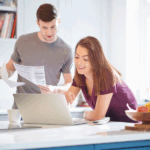 A young man and woman in a kitchen, looking at a laptop. The woman smiles while pointing at the screen, and the man holds papers.