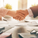 Hands shaking over a desk with documents and a laptop in the background, indicating a business agreement or collaboration.