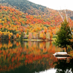 Vibrant autumn foliage reflects in a calm lake, with a small boat docked at the shore and mountains in the background.