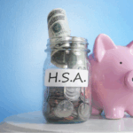 A glass jar labeled "H.S.A." filled with coins and a rolled-up dollar bill, beside a pink piggy bank, against a blue background.