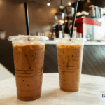Two iced coffee drinks in plastic cups with black straws, one darker and one lighter, placed on a round table in a café setting.