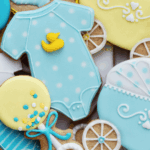 Colorful decorated cookies in various shapes, including baby onesies, strollers, and balloons, primarily in blue and yellow.