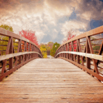 Wooden bridge with a metal railing stretches towards a colorful sky, flanked by trees with autumn foliage in red and orange.