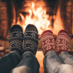 Cozy scene with two pairs of patterned socks by a fireplace, two mugs beside them, flames glowing in the background.