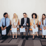 A diverse group of eight job applicants, dressed in business attire, sitting in a row on orange chairs, holding resumes.