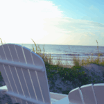 White Adirondack chair facing the ocean, surrounded by tall grass and sandy beach, under a bright blue sky.