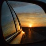 Sunset reflected in a car's side mirror, showing a road with two distant headlights and colorful sky hues of orange and blue.