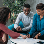 Three people collaborate at a table. A woman in a pink dress points at papers, while a man in a gray sweater and a woman in a blue blouse observe.