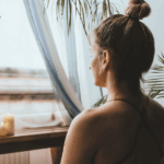 Woman with a bun hairstyle sitting in a serene setting, facing a window with soft light, two candles on a wooden table, plants nearby.