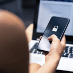 Person entering a passcode on a smartphone with a locked screen, while a laptop with a website is visible in the background.