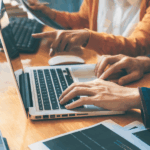 Two hands are typing on a laptop keyboard on a wooden table. One person points at the screen while the other is holding a document.