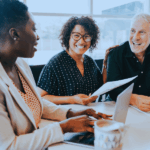 Three people are seated at a table, smiling and engaged in conversation. A woman in a light blazer is typing on a laptop, while another woman holds papers, and a man is laughing. A coffee cup is visible on the table.