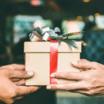 Two hands, one male and one female, are exchanging a small, wrapped gift box with a red ribbon and decorative bow.