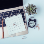 A laptop keyboard with a calendar, clock, succulent plant, paper clips, and a pen arranged on a light blue surface.