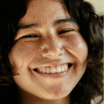 Smiling young person with curly hair and freckles, wearing a nose ring, against a light background.
