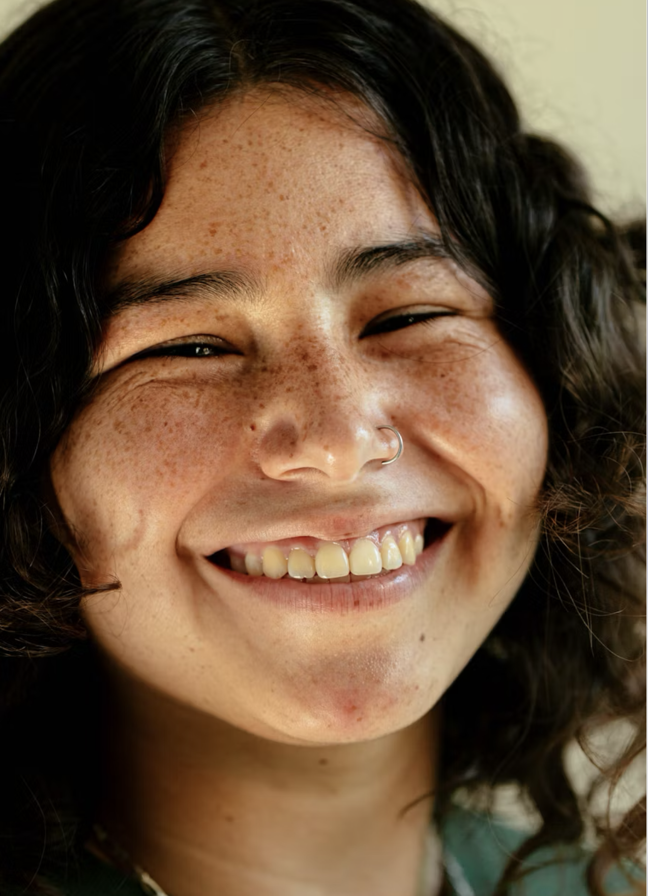 Smiling young person with curly hair and freckles, wearing a nose ring, against a light background.