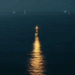 Lone buoy with a bright light in the ocean at night, reflecting golden light on the water surface. Distant lights visible on the horizon.