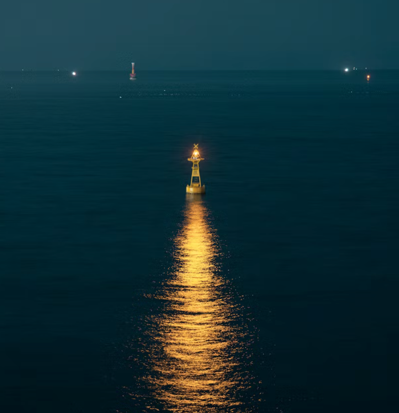 Lone buoy with a bright light in the ocean at night, reflecting golden light on the water surface. Distant lights visible on the horizon.