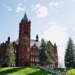 Red brick building with a tall spire, surrounded by green grass and tall trees under a partly cloudy sky.