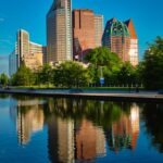 Skyline of tall buildings reflecting in calm water, surrounded by green trees under a clear blue sky.
