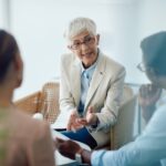 A woman with short gray hair in a beige blazer gestures while speaking to two seated individuals in a bright, modern room.