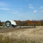 A green barn with a white roof sits in a field under a blue sky with white clouds, surrounded by autumn-colored trees.
