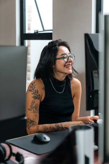 Woman with short dark hair and glasses smiling while sitting at a desk with a computer monitor, wearing a black sleeveless top.