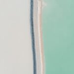 Aerial view of a shoreline with a sandy beach in light beige, bordered by dark gray rocks, and turquoise water.