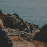 Two elderly individuals sit on a rocky ledge, overlooking a calm body of water at dusk, with a scenic coastline in the background.