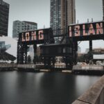 The Long Island sign on a dock structure, surrounded by modern buildings, with a cloudy sky and water in the foreground.