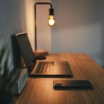 A wooden desk with a laptop, a smartphone, and a lamp casting warm light, with a plant in the background.