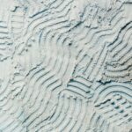 Light blue surface with textured, wavy patterns created by tire or tool marks. Dusty appearance with fine particles.