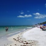 Crowded beach with people swimming and sunbathing under blue umbrellas, against a backdrop of clear blue sky and calm ocean.
