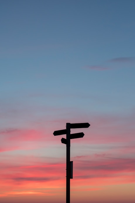 Silhouette of a signpost with multiple arrows against a colorful sunset sky with pink and blue hues.