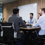 Six professionals engaged in a meeting around a conference table, with a whiteboard in the background.