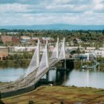 A modern cable-stayed bridge spans a river, surrounded by green trees and city buildings under a partly cloudy sky.