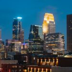 Minneapolis skyline at dusk, featuring illuminated skyscrapers, with the tallest building glowing in golden light.