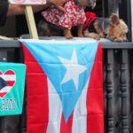 Two small dogs in colorful outfits sit on a balcony with a Puerto Rican flag draped over the railing. A sign reads "Se Regalan Besos."