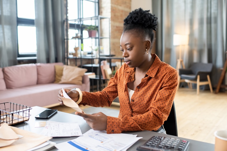 Young Black woman in a brown polka dot shirt sitting at a desk, reviewing documents with a calculator and smartphone nearby.