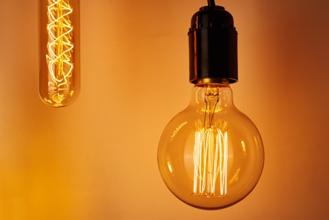 Two light bulbs hang against a warm orange background. One is a vintage-style filament bulb, and the other is a long, thin tube bulb.
