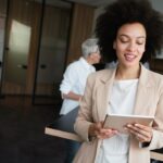 Smiling woman with curly hair in a beige blazer holds a tablet, standing in a modern office with coworkers in the background.