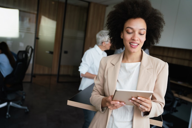 Smiling woman with curly hair in a beige blazer holds a tablet, standing in a modern office with coworkers in the background.