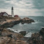 A lighthouse with a red-roofed building stands on rocky cliffs by a choppy sea under a cloudy sky.