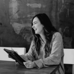 Smiling woman with long hair sits at a wooden table holding a tablet, wearing a cozy gray sweater, against a textured wall.
