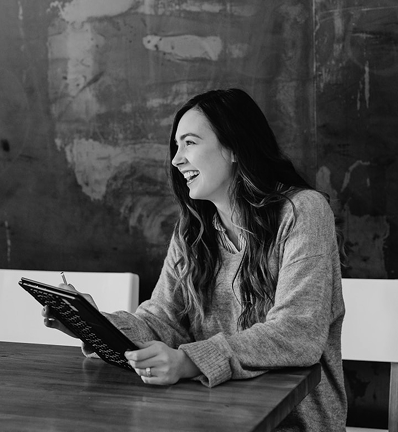 Smiling woman with long hair sits at a wooden table holding a tablet, wearing a cozy gray sweater, against a textured wall.