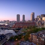 City skyline at dusk featuring tall buildings and waterfront, with marinas and parks visible in the foreground.